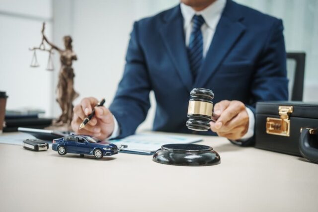 A lawyer in a suit holding a gavel, miniature car, and legal documents nearby.